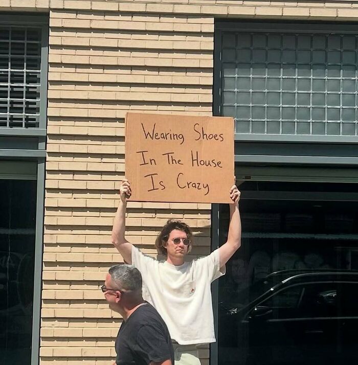 Dude With The Sign holding a cardboard sign about wearing shoes in the house being crazy, outdoors near brick building.