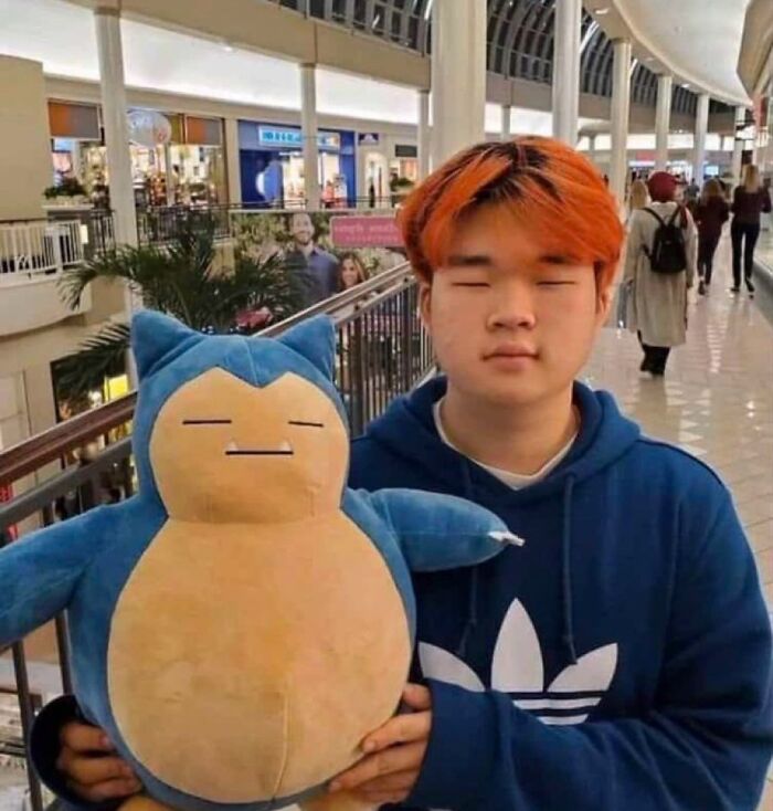 Young man with orange hair wearing a blue hoodie holding a large plush Snorlax in a busy shopping mall hallway.
