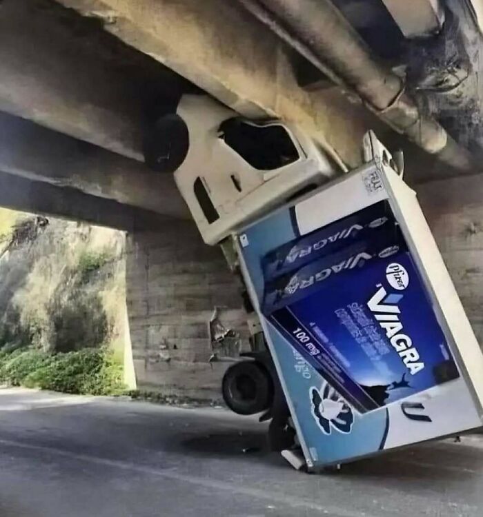 Truck stuck under low bridge with an oversized Viagra box, creating a confusing and absurd scene in a roadside setting.