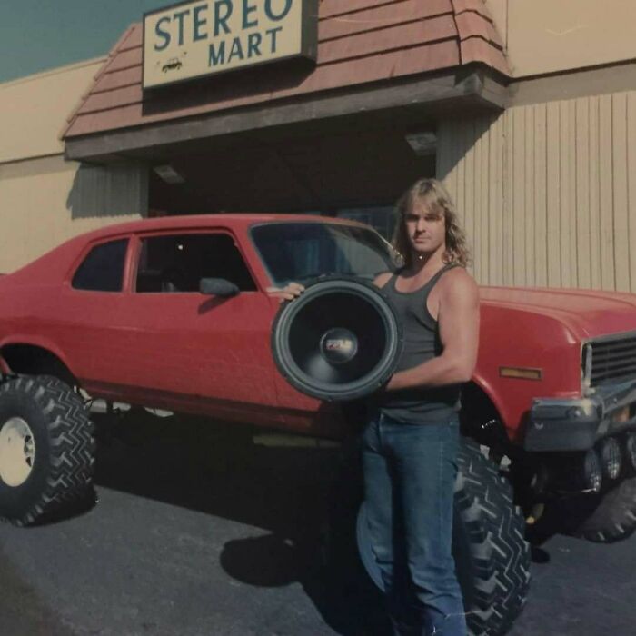 Man embodying old school cool with long hair and tank top holding large speaker in front of lifted red car outside Stereo Mart.