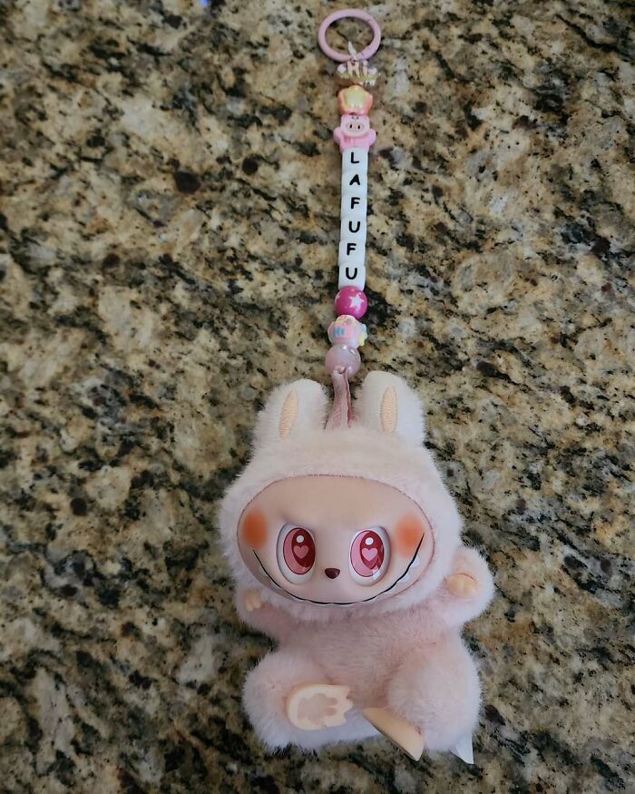 Plush Labubus keychain with creepy smile and large pink eyes hanging on a granite surface, fake collectible toy.