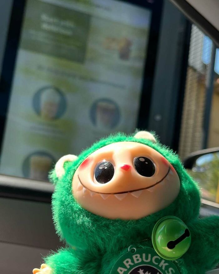Green Labubus fake doll with large eyes and sharp teeth, wearing a Starbucks tag, captured inside a car window.