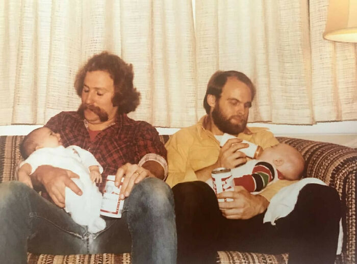 Two dads from the past with classic style, sitting on a couch, each holding a baby and a drink, embodying old school cool.