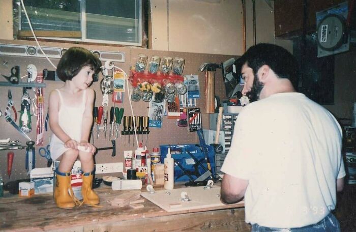 Dad working on a woodworking project in a workshop while a child wearing yellow boots sits on the workbench, old school cool vibe.