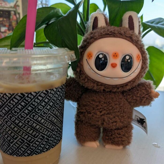Creepy Labubus fake doll with big eyes in a fuzzy brown costume next to a plastic cup of iced coffee inside.