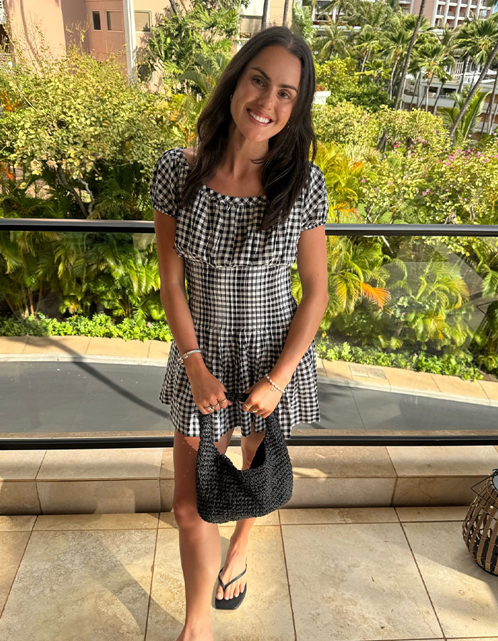 Young influencer in a black and white checkered dress smiling outdoors, amid a lush green background.