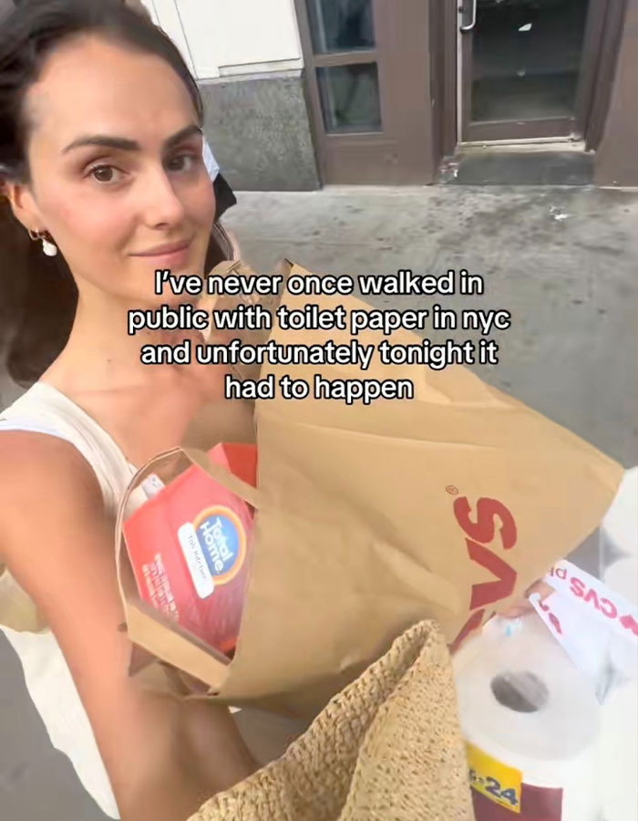 Young woman carrying toilet paper and groceries outside a store, capturing herself on video in a public setting.