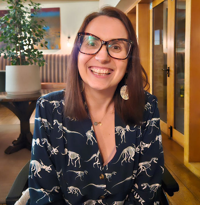 Smiling woman wearing glasses and dinosaur skeleton print shirt, sitting indoors in a cozy room. - 1