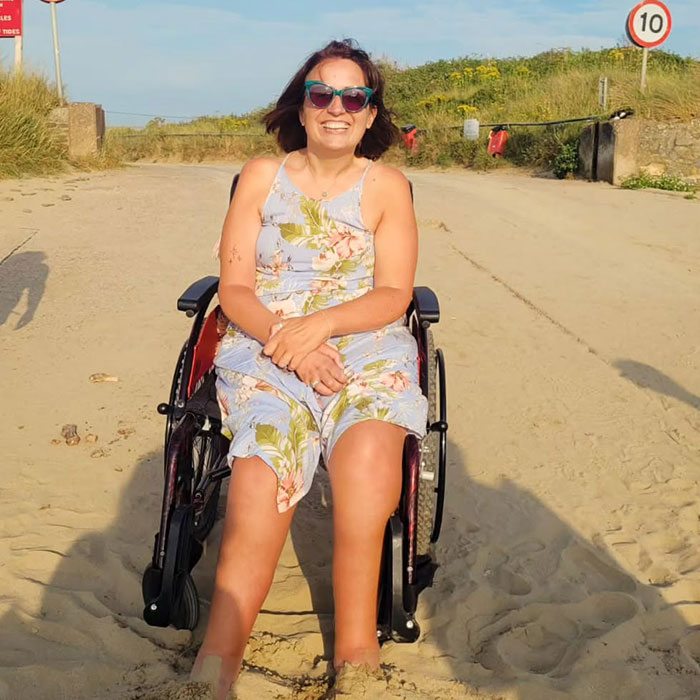 Woman sitting in a wheelchair on a sandy path, wearing sunglasses and a floral dress, smiling in sunlight. - 9