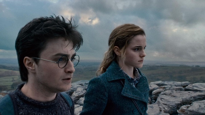Emma Watson standing outdoors with a serious expression beside a man wearing glasses in a cloudy landscape scene. - 9
