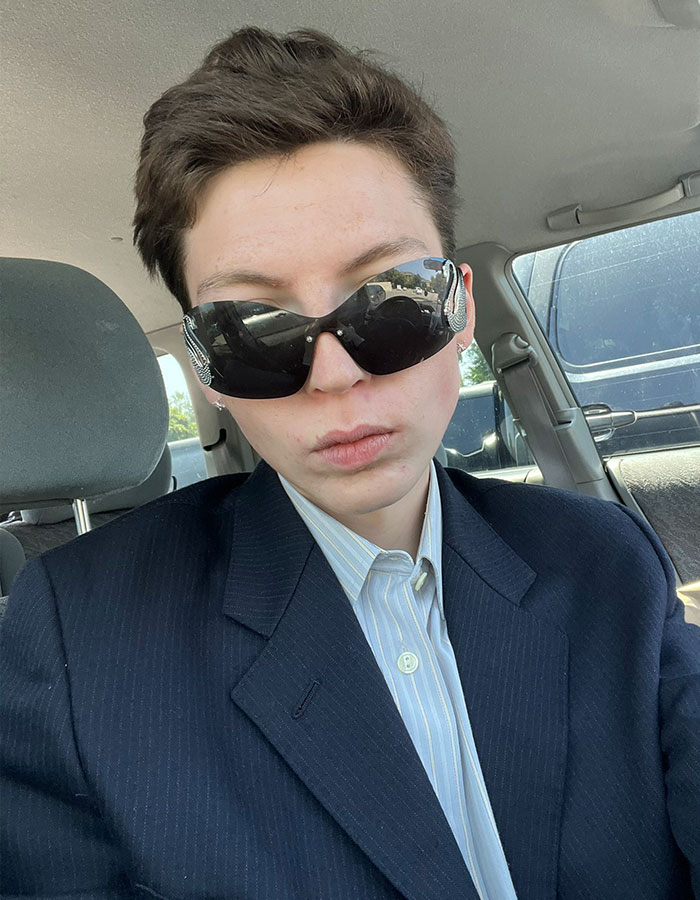 Person with short hair wearing dark sunglasses and a suit jacket sitting inside a car, related to model detransition story. - 2