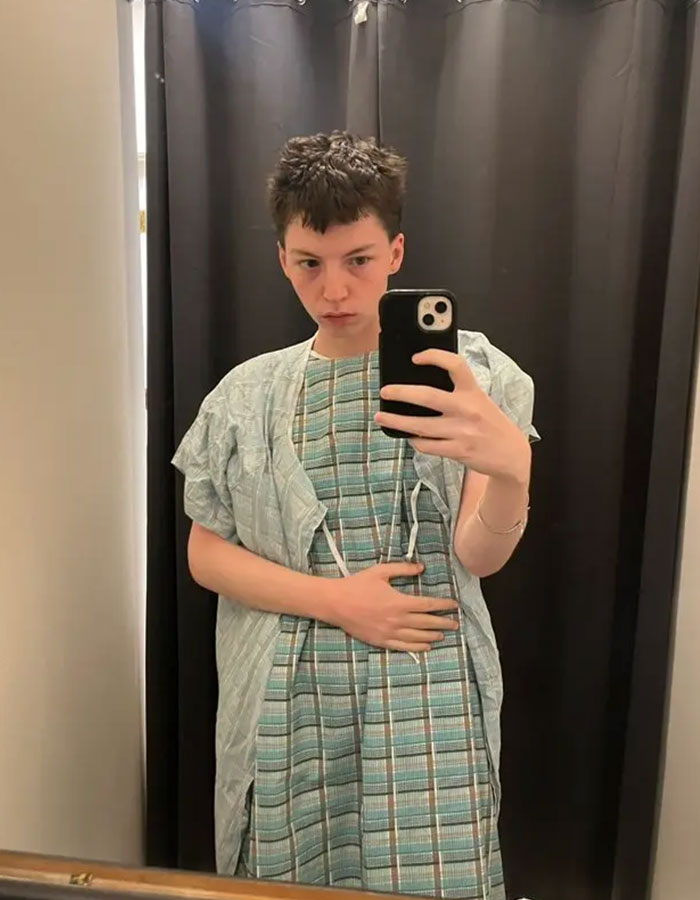 Young person in hospital gown taking a mirror selfie, representing model pushed to transition at 15 now detransitioning. - 6