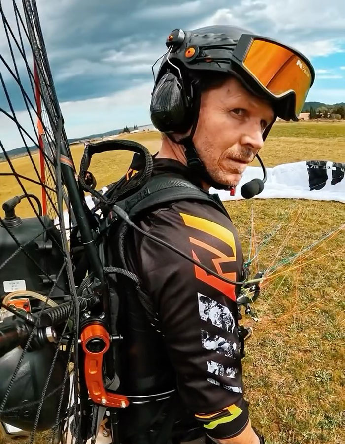 Paramotor pilot wearing helmet and gear preparing for flight on a grassy field under cloudy sky. Paramotor pilot wearing helmet and gear preparing for flight on a grassy field under cloudy sky.