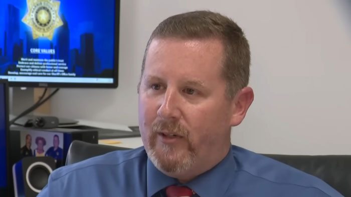Man in a blue shirt and red tie speaking in an office setting with a law enforcement badge on the screen behind him. Man in a blue shirt and red tie speaking in an office setting with a law enforcement badge on the screen behind him.