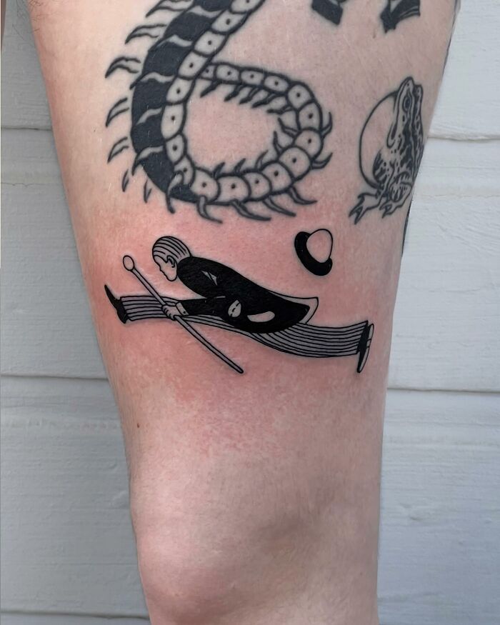 Black ink tattoos on a leg, including a centipede, frog, and a stylized man jumping with a cane and hat.