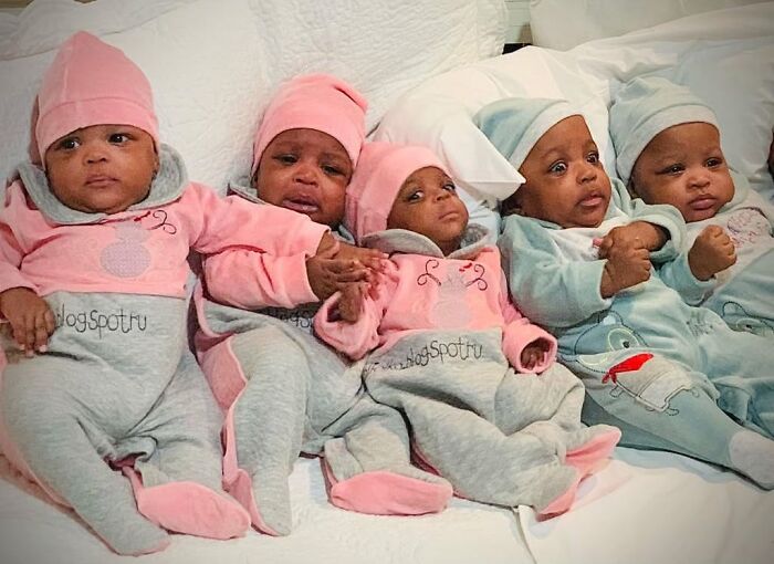 Five of the world&rsquo;s first surviving nonuplets dressed in colorful outfits lying on a white bed.