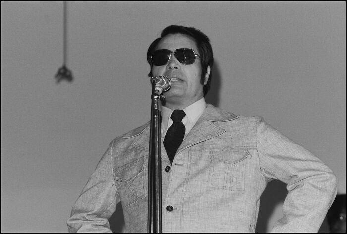 Man wearing sunglasses and a light jacket speaking into a microphone, illustrating unhinged family lore shared by people.