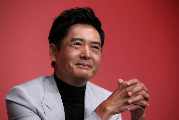 Middle-aged celebrity wearing a light gray suit and black shirt, smiling thoughtfully against a red background.