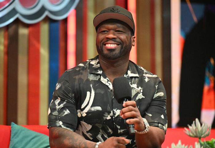 Smiling celebrity holding microphone in casual patterned shirt and cap, exemplifying celebrities choosing not to overspend.