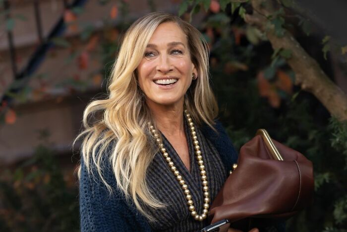 Blonde woman smiling outdoors wearing a dark sweater and gold necklace, holding a brown purse, illustrating celebrities who avoid overspending.