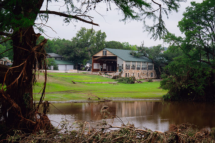 Camp Mystic counselor standing near damaged camp building by the water reflecting the heartbreaking moment returning last surviving kids to parents