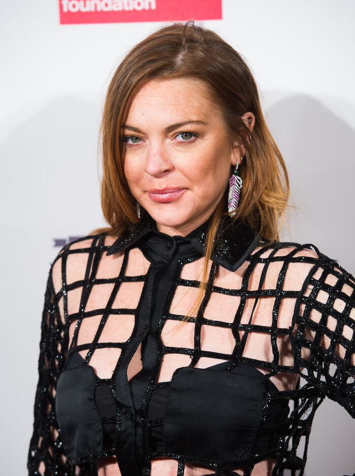 Lindsay Lohan with fresh-faced glow, wearing black mesh outfit and feather earrings at a public event.