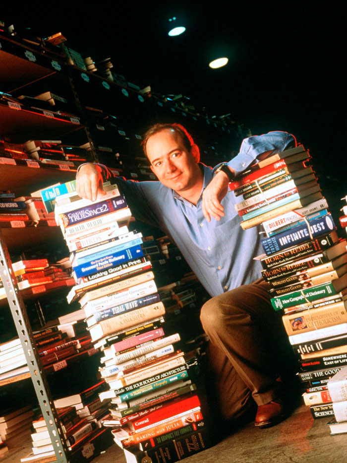 Man resembling Jeff Bezos with hair, leaning against stacks of books in a dimly lit room, ditching his signature bald look.