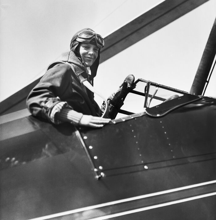 Amelia Earhart in pilot gear sitting in vintage aircraft cockpit with hope for mystery solved from new satellite images.