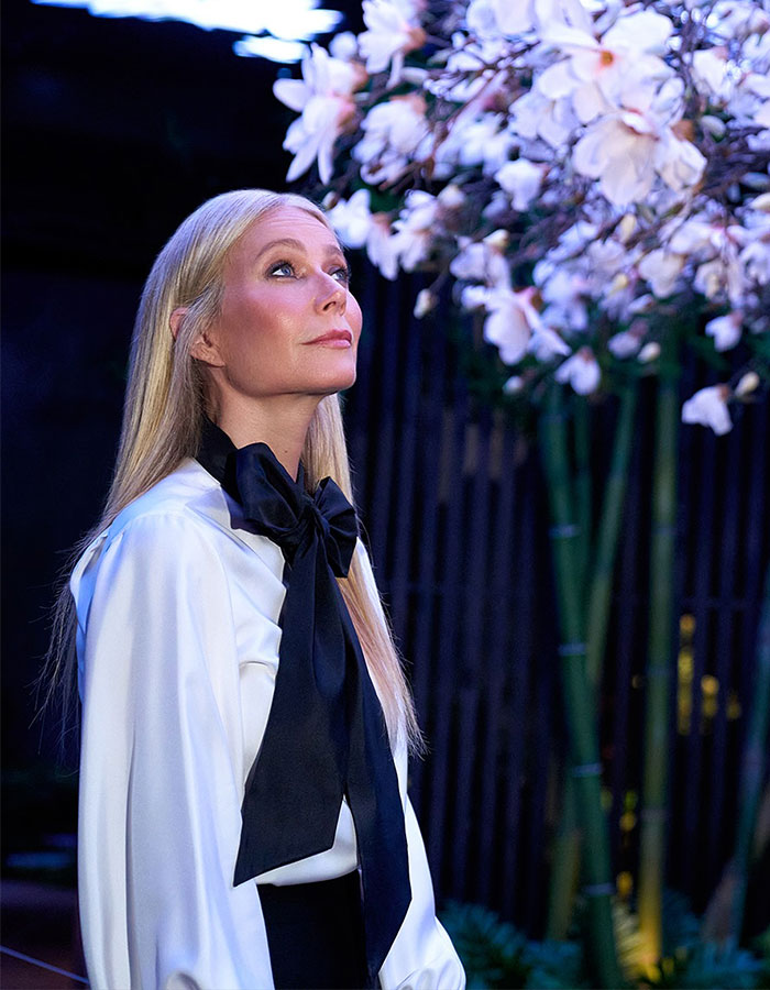 Gwyneth Paltrow in white blouse with black bow, looking up near blooming flowers, related to Coldplay frontman drama. Gwyneth Paltrow in white blouse with black bow, looking up near blooming flowers, related to Coldplay frontman drama.
