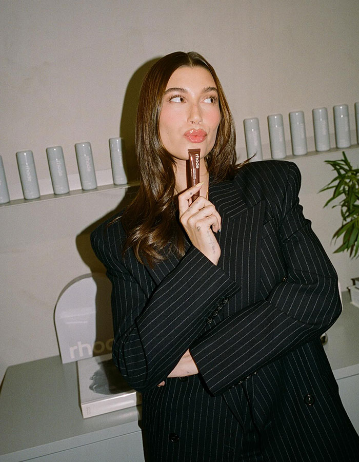 Hailey Bieber posing with Rhode product, wearing a black pinstripe blazer in a minimalist modern setting.