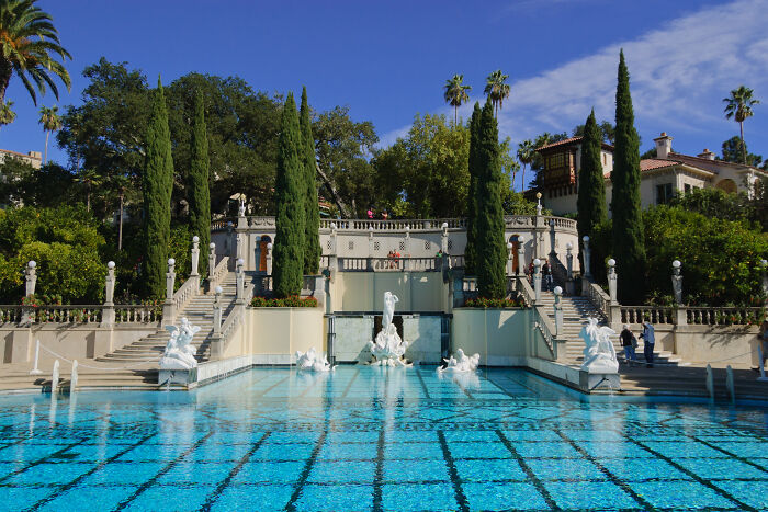 Historic homes with ornate stone statues and vibrant blue pool surrounded by tall trees under a clear sky.
