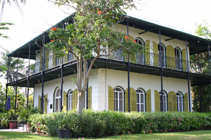 Two-story historic home with wraparound balcony, arched windows, and green shutters surrounded by lush greenery.