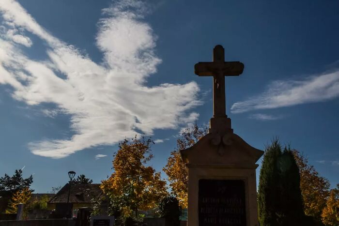 Silhouette of a cross monument against a blue sky with clouds resembling a bird, captured by a street photographer.