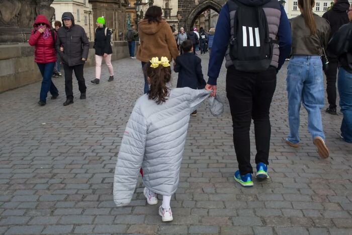Child wearing oversized jacket walking with adult on cobblestone street, a serendipitous moment captured by street photographer.