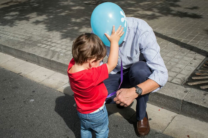 Child holding a blue balloon while a man kneels on the sidewalk, capturing a serendipitous moment in street photography.