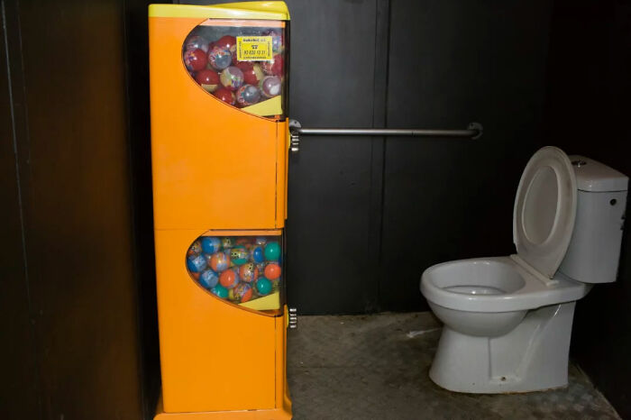 Yellow capsule toy vending machine next to a white toilet in a dimly lit room, capturing a serendipitous moment in everyday life.