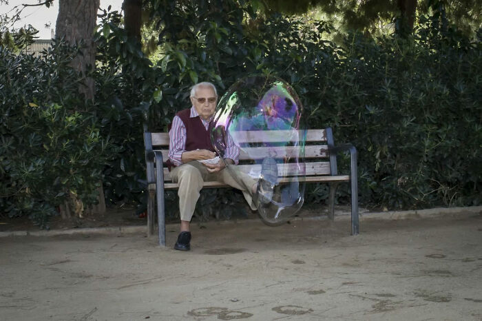 Elderly man sitting on park bench with a giant bubble floating nearby, captured in a street photography moment.