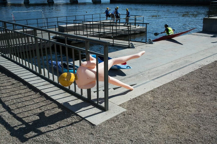 Inflatable doll caught awkwardly on railing near waterfront with kayakers and people walking by in the background.