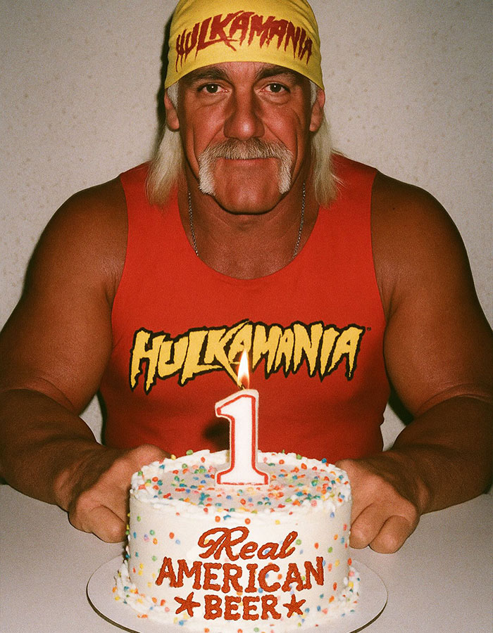 Hulk Hogan wearing red Hulkamania shirt and yellow bandana, holding a cake with a lit number one candle. - 2
