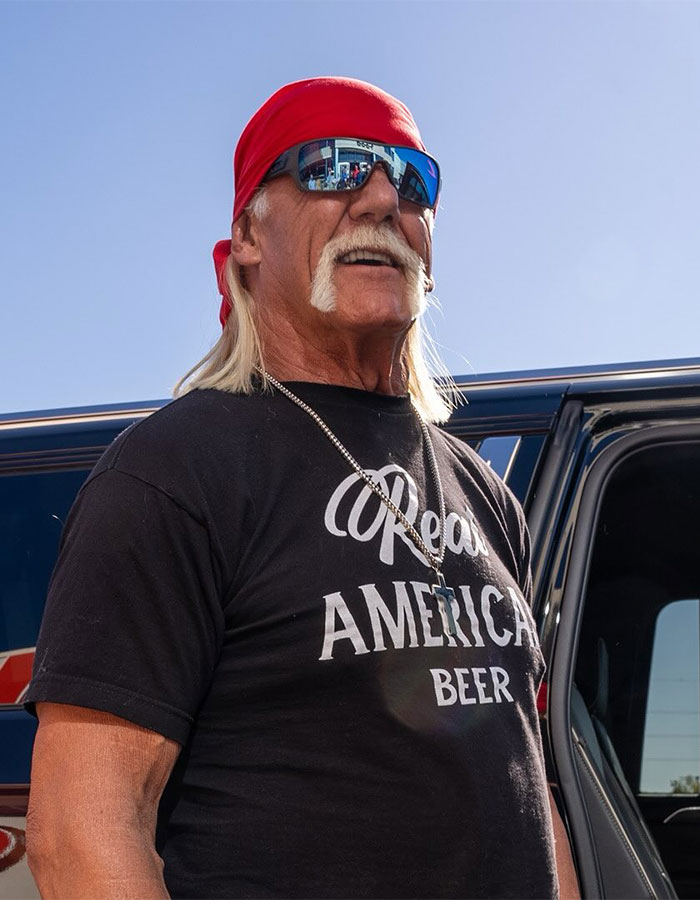 WWE legend Hulk Hogan wearing sunglasses and a red bandana, standing outside near a vehicle on a sunny day. - 3
