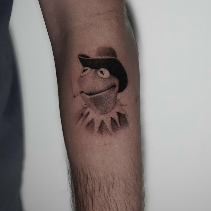Black and gray tattoo of a frog wearing a hat and smoking on a person's forearm, showcasing one of the best tattoos.