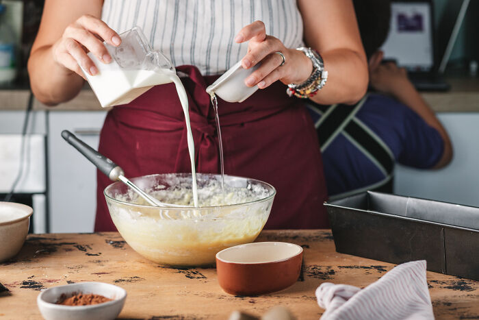 Person wearing apron pouring milk and another liquid into a bowl, baking childhood cake as a family remedy after hospital trip. - 5
