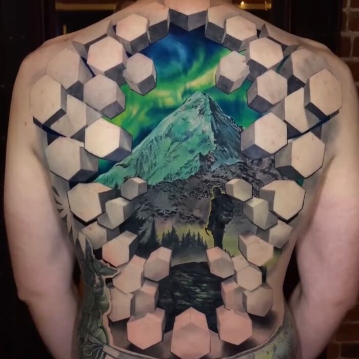 3D tattoo on back by Jesse Rix featuring hexagonal shapes framing a mountain and a person looking at a portal landscape.