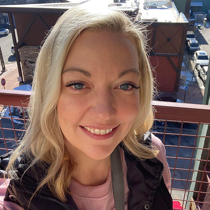 Smiling woman with blonde hair in a casual outfit, pictured outdoors on a sunny day with urban background.