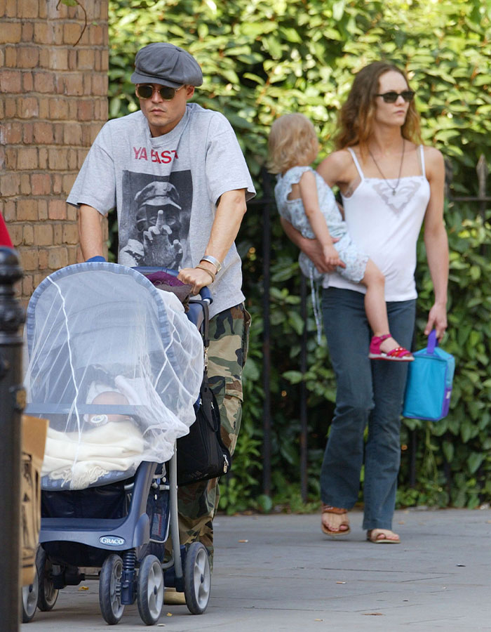 Johnny Depp wearing hat and sunglasses pushing stroller outdoors with a woman holding a child in the background.