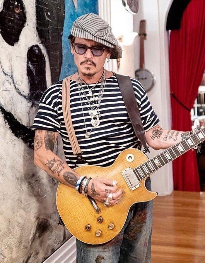 Johnny Depp playing guitar indoors, wearing a striped shirt and hat, showing tattoos and layered necklaces.