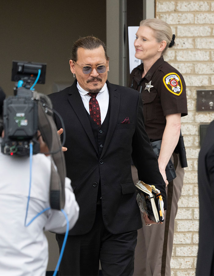 Johnny Depp dressed in black suit and sunglasses, holding documents, as a police officer stands nearby outside a building.