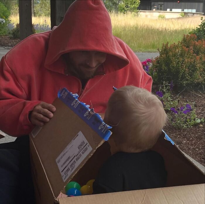 Man in red hoodie smiling at child playing inside cardboard box outdoors, reflecting on marriage and mental struggles.