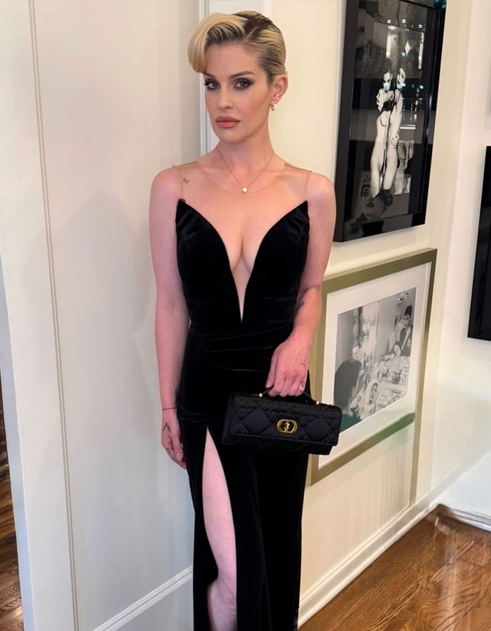 Kelly Osbourne in a black velvet 90s glam dress with a thigh-high slit, holding a black quilted purse indoors. Kelly Osbourne in a black velvet 90s glam dress with a thigh-high slit, holding a black quilted purse indoors.
