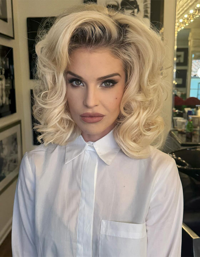 Kelly Osbourne styled in 90s glam fashion with voluminous curly blonde hair and bold makeup in a white collared shirt. Kelly Osbourne styled in 90s glam fashion with voluminous curly blonde hair and bold makeup in a white collared shirt.
